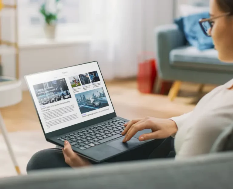 A person sits on a couch using a laptop, viewing an online news website with articles and images displayed on the screen.