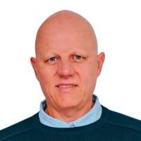 A middle-aged person with a bald head is wearing a blue collared shirt and dark sweater, facing the camera against a plain white background.