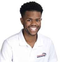 A man with short curly hair and a beard smiles at the camera, wearing a white collared shirt with a logo on it, in front of a plain white background.