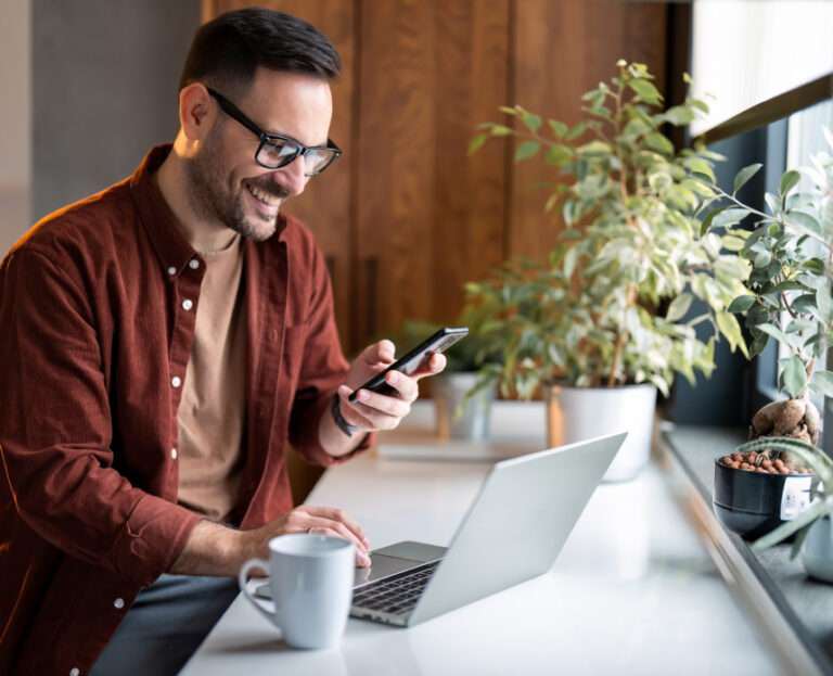 A man wearing glasses sits at a desk with a laptop, holding a smartphone and smiling, with seo software on his screen, a coffee mug, and potted plants nearby.