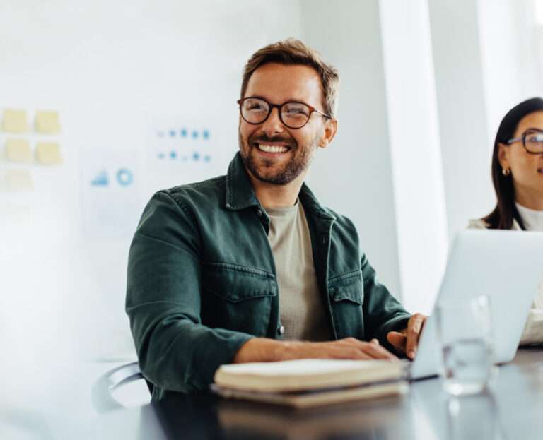A man wearing glasses and a green jacket sits at a table with a laptop and notebook, smiling as he discusses seo software. Another person is partially visible beside him.