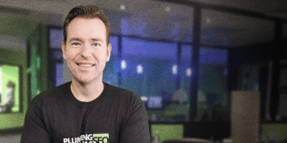 A man wearing a black "PLUMBING SEO" t-shirt smiles while standing in an office with glass walls and green accents.