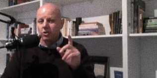 A bald man speaks into a microphone, gesturing with his hand, while seated in front of bookshelves filled with books and various items.