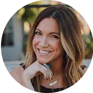 A woman with shoulder-length brown hair smiles at the camera, resting her chin on her hand. She wears a necklace and a sleeveless top.
