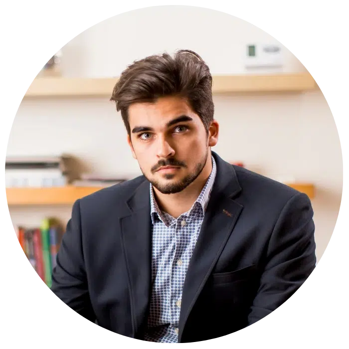 A man with dark hair and a beard, wearing a blazer and checked shirt, sits in front of shelves with books and decor.