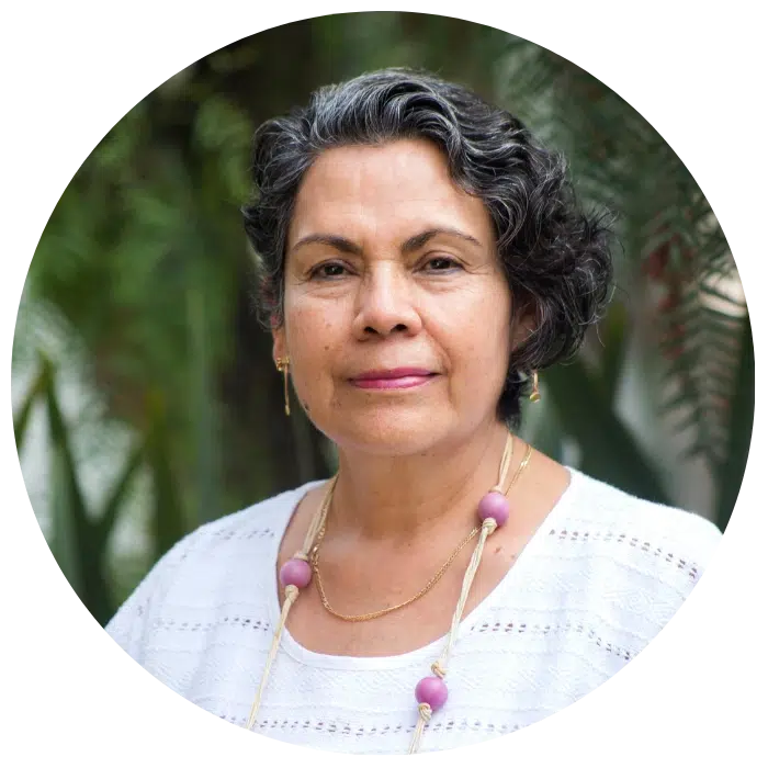 A middle-aged woman with short curly gray hair, wearing a white top and a necklace with large beads, stands outdoors with greenery in the background.