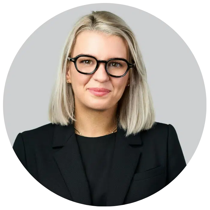 A woman with shoulder-length blonde hair and black-rimmed glasses, wearing a black blazer, poses against a plain light gray background.
