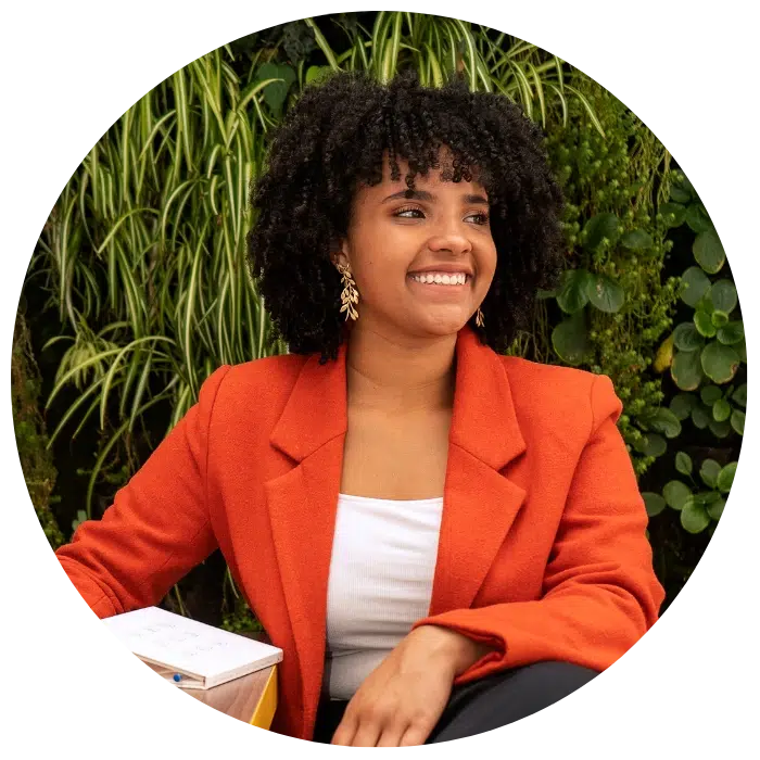 A woman with curly hair in an orange blazer and white top sits and smiles, with greenery in the background.