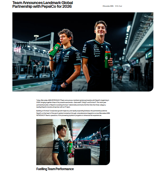 Two Mercedes-AMG PETRONAS F1 team members stand in team uniforms holding PepsiCo drinks at a racetrack, with a racing car and pit lane in the background.